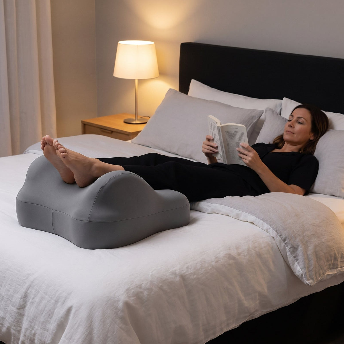 Woman reading in bed with her legs raised on the gray LegLoft leg rest pillow