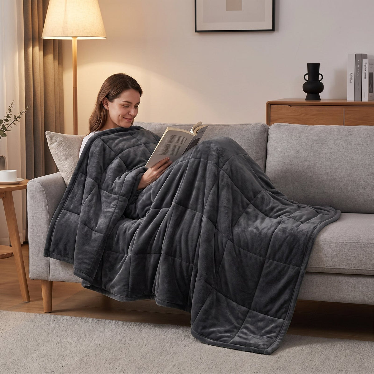 Woman reading on a light gray sofa under the dark gray GravityRest weighted blanket